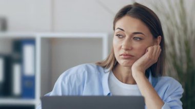 Genç bir kadın parlak bir ofiste bir masada oturuyor, çenesini elinde dinlendiriyor, dizüstü bilgisayarına bakarken sıkılmış görünüyor. Mekan düzenli ve minimalistik..
