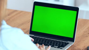 Bir insan, düz yeşil bir ekran gösteren dizüstü bilgisayarına odaklanır. Dekor, ahşap masası ve minimum dağınıklığı olan rahat bir çalışma alanı, verimli bir atmosfer yaratıyor..