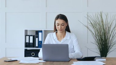Odaklanmış genç bir kadın masasında oturuyor, kağıtların arasında bir dizüstü bilgisayar ve modern ofis ortamını gösteren organize bir raf yazıyor. Atmosfer sakin ve profesyonel..