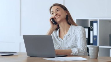 Parlak ofis ortamında telefonuyla konuşan bir kadın gülümsüyor. Ofis malzemeleri ve kahve fincanı ile çevrili bir dizüstü bilgisayarla bir masada oturuyor..