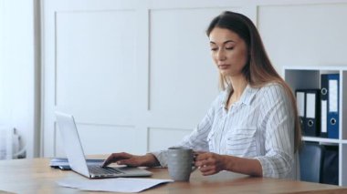Bir kadın elinde bir kupa kahve tutarken dizüstü bilgisayarına odaklanmış bir şekilde ahşap masada oturuyor. Ofis iyi aydınlatılmış, organize klasörler ve üretkenliğe olanak sağlayan sakin bir atmosferi var..