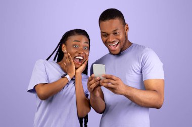 Heyecanlı siyah çift cep telefonu ekranına bakıyor, yeni bir uygulama kullanıyor, menekşe turuncu stüdyo arka planı. Neşeli Afro-Amerikan erkek ve kadın akıllı telefon kullanıyorlar, eğlence konsepti.