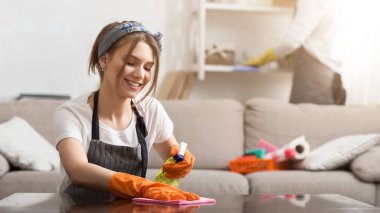 Milenyum çifti birlikte bahar temizliği yaparken, kadın, erkek arkadaşı tozdan rafları yıkarken bez ve spreyle masayı siliyor.