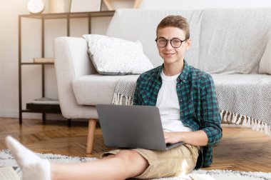 Gözlüklü, ev ödevi yapan, kitap ve dizüstü bilgisayar kullanarak yerde oturan neşeli bir genç.