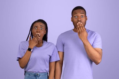 Omg. Korkmuş Afro-Amerikan çiftin portresi ağızlarını eliyle kapatıyor, şok olmuş kadın ve adam mor stüdyo arka planında izole bir şekilde kameraya bakıyor. İnsani duygular, yüz ifadesi