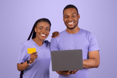 Çevrimiçi Bankacılık ve E-Ticaret Konsepti. Mutlu siyah çift, elinde bankamatik kartı tutan neşeli bir kadın menekşe mor stüdyo duvarına izole edilmiş, bilgisayarla online alışveriş yapıyor.