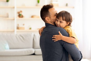 Sevimli küçük çocuk, sevgi dolu babasıyla eğleniyor, sevgi dolu bir adam, sevgili oğlunu kucaklıyor ve öpüyor, evde el ele tutuşuyor.