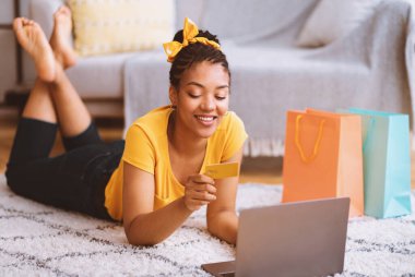 Çevrimiçi İşlem Kavramı. Gülümseyen siyah kadın dizüstü bilgisayar ve kredi kartı kullanıyor, internetten alışveriş siparişi veriyor, zemin halısında yatıyor, banka kartı numarasını kontrol ediyor ve CVV güvenlik kodu yazıyor.