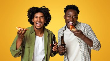 Mens Leisure Concept. Two Black Guys Holding Beer Bottles And Eating Pizza, Cheerful African American Male Friends Having Fun Together, Standing Over Beige Studio Background And Looking At Camera