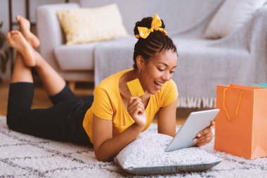 Heyecanlı Afrikalı kız kredi kartı gösteriyor, tablet kullanıyor, yerde yatıyor.