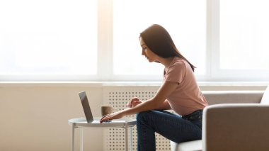 Ev penceresinin yanındaki koltukta oturan dizüstü bilgisayarda çalışan serbest çalışan bir kadın. Modern yaşam tarzı ve uzak kariyer. Yan Görünüm