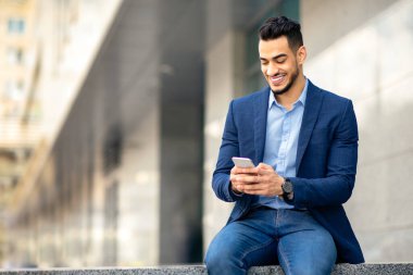 İş yeri binasının yanında oturan resmi elbiseli başarılı bir Arap adam akıllı telefon ve gülümseme kullanıyor, kopyalama alanı kullanıyor. Orta Doğulu işadamı iş başvurusunu cep telefonuyla kullanıyor