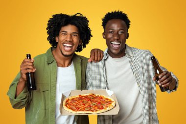 Erkek Partisi. Bira ve pizza kutusundaki iki mutlu siyah arkadaş, bej rengi stüdyoda poz veriyorlar, Afro-amerikan erkeklerinin lezzetli atıştırmalıkları ve içkileri ile portresi,