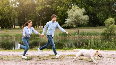 Aktif Hayvan Konsepti. Gülen çift göl kenarındaki parkta köpeğini kovalıyor. Kopya alanı, tam boy.