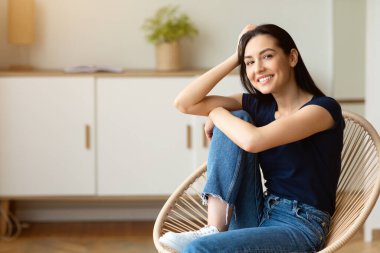 Modern sandalyede oturan rahat bir kız kameraya gülümsüyor, evde rahat bir hafta sonunun tadını çıkarıyor. Boşluğu Kopyala
