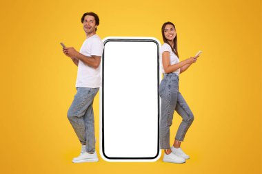 Modern İletişim. Mutlu Neşeli Çiftin Tüm Uzunluğu Büyük Akıllı Telefona yaslanıp Beyaz Boş Ekran ile İki Cep Telefonu Kullanarak, Sosyal medyanın Turuncu Arkaplanda Duruşunda Sohbet Etmesi