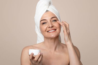 Yaşlanma önleyici cilt bakımı. Mutlu olgun bir kadın yüzüne nemlendirici krem sürüyor, banyodan sonra kafasında havluyla poz veriyor.