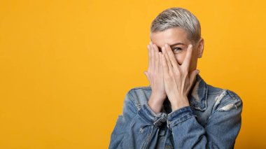 Çekingen, utangaç, olgun bir kadının portresi. Elleriyle yüzünü kapatıyor ve parmaklarıyla dikizliyor. Sarı arka planda duruyor.