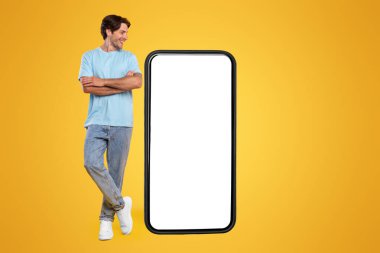 Neşeli, güler yüzlü, elleri çapraz duran koca cep telefonuna, boş beyaz ekran şablonuna, turuncu stüdyo duvarına bakan sıradan bir adam. Uygulama ya da web sitesi için, ücretsiz kopyalama alanı, tam vücut uzunluğu