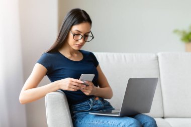 Dizüstü bilgisayarlı kız cep telefonu kullanıyor. İnterneti tarıyor. Evdeki koltukta oturuyor. Mobil Uygulama Konsepti