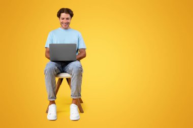 Sırıtkan sakallı adamın portresi dizüstü bilgisayar kullanarak, haberleri okuyarak, video ya da film izleyerek, çalışarak ya da uzaktan kumandayla çalışarak, sarı stüdyo arka planında, ön dikey çekimde izole edilmiş,