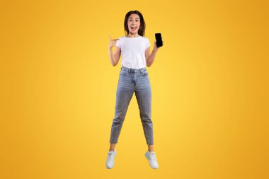 Cool Mobil Uygulama. Heyecanlı sıradan bir kadın boş ekranlı akıllı telefonu işaret ediyor ve sarı stüdyo arka planında zıplıyor. Model, reklam, web sitesi veya uygulama tasarımı için boş alan