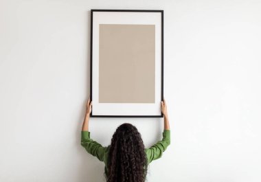 Unrecognizable Female Hanging Blank Poster In Frame On White Wall Standing Back To Camera Indoors. Modern Art And Decor For Domestic Interior Concept. Mockup