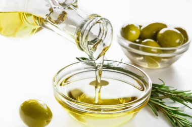 Şişeden dökülen zeytinyağı, kaselerde taze zeytin ve beyaz arka planda biberiye sprigs