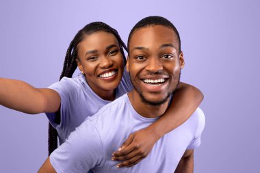 Eğleniyorum. Mutlu siyah çift portresi konuşan selfie, neşeli kadın erkek arkadaşlarının sırtına biniyor, mor stüdyo duvarının üzerinde izole bir şekilde duran kameraya bakıyor.