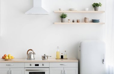 Minimalist mutfağı ve İskandinav iç mimarisi olan modern bir daire. Sobalı beyaz mobilyalar, çaydanlık ve çatal bıçak. Tabakta meyveler, tabakta tabaklar, saksıda bitkiler, aydınlık duvar.