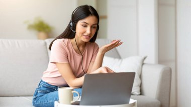 Kulaklıklı kız, evdeki koltukta oturan dizüstü bilgisayarla video görüşmesi yapıyor. Uzaktan Çalışma Konsepti. Seçici Odaklanma