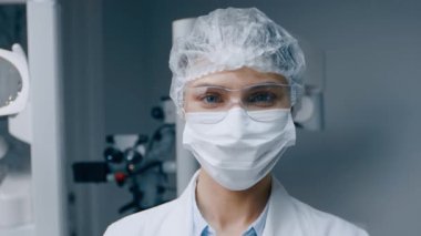 Laboratuvar önlüğü ve koruyucu giysileri içinde bir dişçi kadın modern bir klinikte bakıma hazır bekliyor. Ayarlarda gelişmiş diş donanımı ve hastanın güvenliği ve sağlığı üzerinde duruluyor.