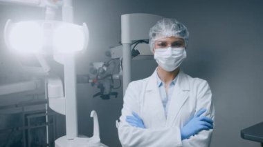 Laboratuvar önlüğü ve koruyucu giysi giyen bir dişçi kadın modern bir diş kliniğinde, kollarını kavuşturmuş, hastaya bakımı için hazır bekliyor. Oda gelişmiş tıbbi aletlerle donatılmıştır..
