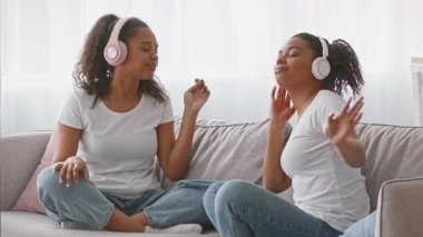 İki Afrikalı Amerikalı kız kardeş evdeki bir kanepede oturuyorlar, kulaklık takıp müzik dinliyorlar. Neşeli ifadeleri yakın bir bağı ve kardeşlik anlarını yansıtıyor..