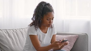 Genç bir Afrikalı Amerikalı kız rahat bir kanepede tek başına oturuyor ve akıllı telefonuna odaklanıyor. Güneş ışığı perdelerin arasından süzülüp sıcak ve davetkar bir atmosfer yaratır..