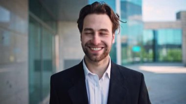 Bin yıllık bir adam şık bir şirket binasının önünde takım elbise giymiş ve gülümsüyor. İfadesi, girişimci ve yönetici olarak kariyerinde karşılaştığı zorluklar arasında iyimserlik taşıyor..