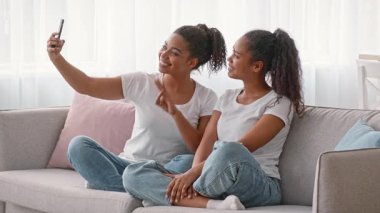 İki Afrikalı Amerikalı kız kardeş bir kanepeye yakın otururlar, gülümserler ve selfie çekerler. Aralarındaki bağ sevgiyi ve mutluluğu yansıtıyor. Sıcaklıkla dolu sıcak bir yuva ortamında..
