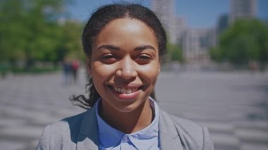 Genç bir Afrikalı Amerikalı kadın, profesyonel giysiler içinde güvenle gülümsüyor. Canlı bir şehir parkında duruyor, güzel havanın ve etrafındaki hayat dolu atmosferin tadını çıkarıyor..