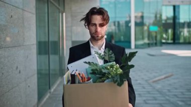 Takım elbiseli bir milenyum adamı bir kutu kişisel eşyayla birlikte bir şirket binasını terk eder. Ofis malzemeleri ve bir bitki taşıdığı için kasvetli görünüyor. Bu da işini kaybettiğini gösteriyor..