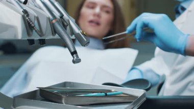 Bir dişçi, çağdaş bir diş kliniğinde hastasının dişlerini muayene ediyor. Hasta dişçi koltuğunda otururken, aletler yakındaki bir tepside düzenlenirken, ağız sağlığı hizmetlerine odaklanır..