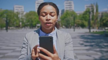 Profesyonel bir Afro-Amerikan kadın dışarıda duruyor, akıllı telefonuna odaklanmış. Takım elbise giyip, şehir yeşilliğine karşı önemli bir görev üstlenirken kendine güveni tamdır..