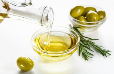 Kavanozdan dökülen zeytinyağı, kaselerde taze zeytin ve beyaz arka planda biberiye sprigs