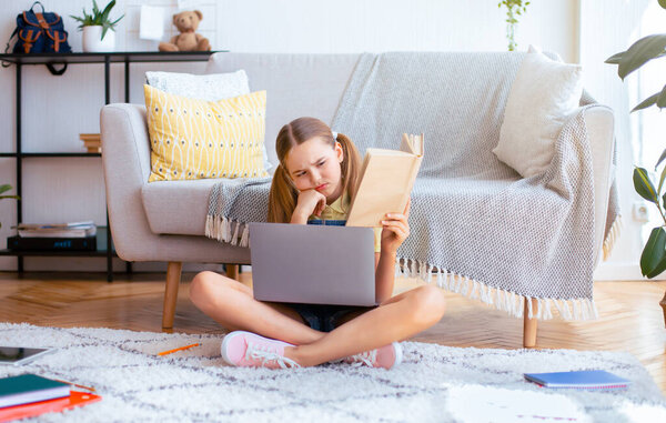 Портрет несчастной школьницы, сидящей с ноутбуком на полу, держащей в руках бумажную книгу