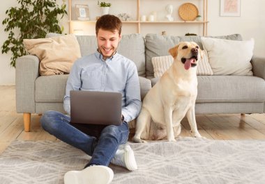 Çalışan Ev Konsepti. Oturma odasında Labrador 'uyla halıda oturmuş dizüstü bilgisayar kullanan gülümseyen bir adamın portresi. İnternette serbest çalışan mutlu bir adam, yerde telgraf çekiyor.