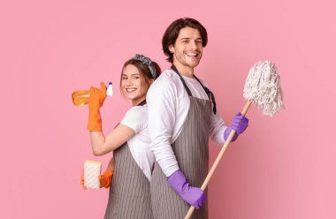 Profesyonel Temizlik Servisi. Gülümseyen adam ve kadının portresi. Ev aletleri ellerinde. Pembe stüdyonun arka planında sırt sırta duruyorlar.