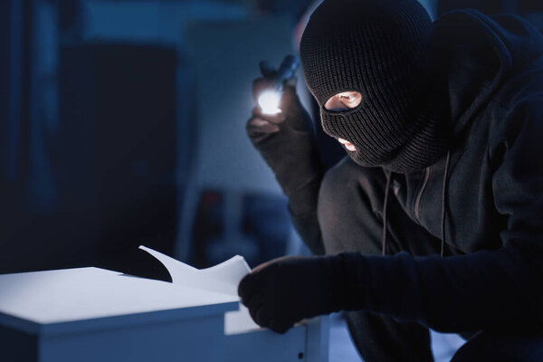 Industrial Espionage. Masked intruder holding and reading confidential documents using flashlight at night, copy space