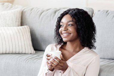 Ev Konforu. Neşeli genç Afrikalı Amerikalı kadın elinde bir fincan çayla yerde dinleniyor, kahve kupası tutuyor, rahat bir oturma odasında dinleniyor, hafta sonunun keyfini çıkarıyor, izin gününün tadını çıkarıyor, fotokopisini çekiyor.