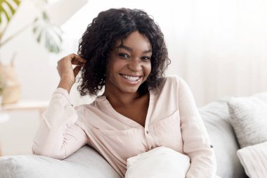 Yastıkla koltukta oturan, rahat bir oturma odasında dinlenen ve kameraya gülümseyen mutlu siyah kadın.