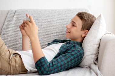 Evde cep telefonuyla oyun oynayan mutlu bir gencin yan görünüşü, kanepede uzanması, kopyalama alanı.