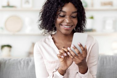 Neşeli siyah kız evde akıllı telefon kullanıyor, arkadaşlarıyla internette mesajlaşıyor, rahat koltukta dinleniyor.